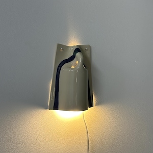 May include: A wall-mounted ceramic light fixture with a glossy off-white finish and a dark blue vertical stripe. The sconce features a curved, folded design that emits a warm, glowing light from both the top and bottom openings, with a clear cord.