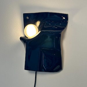 Puede incluir: Un aplique de pared con una base de cer&aacute;mica azul marino oscuro brillante de forma irregular y plegada. Una bombilla blanca redonda y brillante est&aacute; colocada dentro de una hendidura curva en el lado izquierdo de la superficie oscura y reflectante.