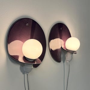 K&ouml;nnte beinhalten: Zwei Wandleuchten mit kreisf&ouml;rmigen, dunkelvioletten, reflektierenden R&uuml;ckplatten in gl&auml;nzender Ausf&uuml;hrung. Jede Leuchte verf&uuml;gt &uuml;ber eine wei&szlig;e, kugelf&ouml;rmige Gl&uuml;hbirne an einer wei&szlig;en Fassung und ein d&uuml;nnes, helles Stromkabel.