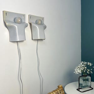 May include: Two matching wall-mounted light fixtures with a smooth, light gray ceramic finish. Each fixture features a rectangular top section with a circular cutout holding a round, frosted glass light bulb. A thin, clear power cord hangs down.