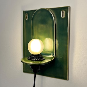 May include: A wall-mounted ceramic sconce featuring a glossy dark green finish with a rectangular backplate and a curved shelf. A small, frosted spherical light bulb sits on the shelf, connected to a black cord extending downward from the base.