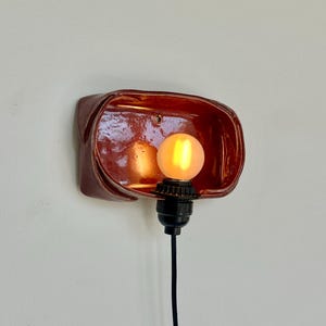 May include: A wall-mounted, reddish-brown ceramic light fixture with a glossy finish. The fixture has a rectangular shape with rounded edges, housing a lit, round, yellow light bulb. A black cord extends downward from the bulb.