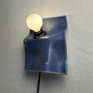 May include: A wall-mounted lamp featuring a folded, deep blue ceramic square plate with a smooth, glossy finish. A black socket holds a single, glowing round white light bulb. A thin black power cord extends downward from the base of the fixture.