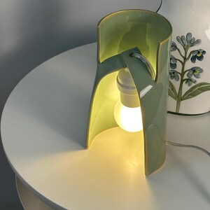 May include: A sage green ceramic table lamp with a curved, open-sided cylindrical structure. The lamp features a central white light bulb socket and a glowing bulb, with a thin white power cord extending from the base of the ceramic body.