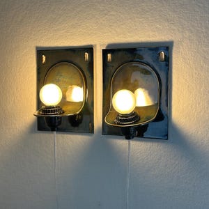 May include: Two wall-mounted sconces with a dark blue and gold finish. Each sconce features a rounded light fixture with a spherical bulb. The sconces are rectangular with a curved top and a small shelf to hold the bulb. The sconces are wired.