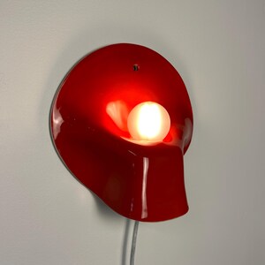 May include: A glossy red wall lamp with a curved, organic shape and a single circular opening at the top. A round, frosted white light bulb is positioned in the center, emitting a warm glow. A thin, light-colored power cord hangs from the bottom.