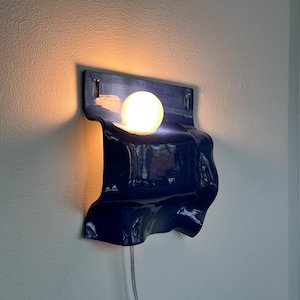 May include: A modern, navy blue wall sconce with a unique, flowing design. The sconce features a spherical light bulb that emits a warm glow. A clear cord extends from the bottom. The fixture is mounted on a white wall.