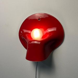 May include: A glossy red wall lamp with a curved, organic shape and a central circular opening. A single glowing white spherical light bulb is positioned within the center of the red fixture. A thin black power cord hangs from the bottom of the lamp.
