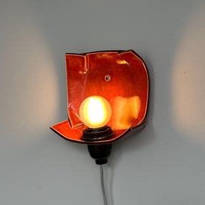 May include: A wall-mounted light fixture with a warm, orange-red shade. The fixture has a black base and a round, glowing light bulb. The fixture is mounted on a light gray wall.