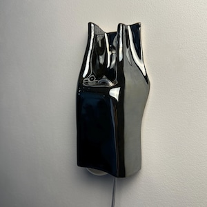 May include: A wall-mounted light fixture with a glossy, reflective black ceramic finish. The object features an elongated, organic, and undulating form with a smooth surface. A thin, light-colored electrical cord extends from the bottom edge.