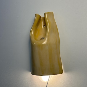 Pu&ograve; includere: Una lampada da parete in ceramica con una finitura lucida color giallo senape. L'oggetto presenta una forma organica, piegata e asimmetrica con un centro cavo. Una singola lampadina rotonda &egrave; visibile in basso, emettendo una luce calda.