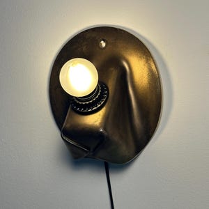 May include: A wall-mounted lamp featuring a circular, metallic bronze-toned backplate with a draped, fabric-like texture. A single round, glowing light bulb is secured in a black socket, and a thin black power cord extends downward from the base.