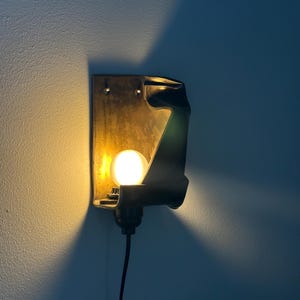 May include: A wall-mounted sconce featuring a dark, curved metal housing with a textured finish. A single round, glowing light bulb is positioned within the structure, casting a warm yellow light against the wall and creating a distinct shadow.