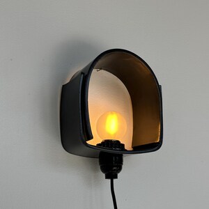 Puede incluir: Una l&aacute;mpara de pared negra con una pantalla curva en forma de visera. La l&aacute;mpara alberga una bombilla redonda e iluminada. Un cable negro se extiende hacia abajo desde la base, y la l&aacute;mpara est&aacute; montada en una pared de color claro.