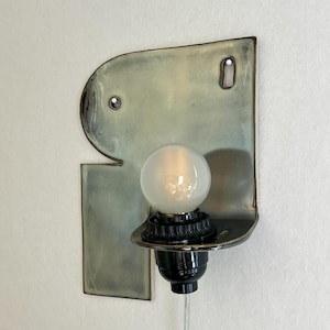 May include: A wall-mounted light fixture featuring a flat, irregularly shaped ceramic backplate with a glossy, pale green and grey glaze. A black socket holds a single round, frosted white light bulb, with a clear power cord extending downward.