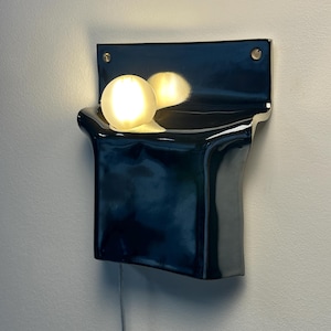 Peut inclure: Une applique murale en c&eacute;ramique noire avec une finition brillante et une plaque arri&egrave;re rectangulaire dot&eacute;e de deux trous de fixation. Le luminaire comprend une ampoule blanche sph&eacute;rique qui &eacute;met une lueur chaude et se refl&egrave;te sur la surface.