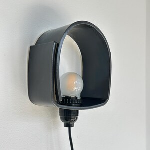 Puede incluir: Una l&aacute;mpara de pared negra con una pantalla curva en forma de arco. La l&aacute;mpara alberga una bombilla y est&aacute; conectada a un cable el&eacute;ctrico negro. La luz est&aacute; montada en una pared blanca.