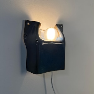 May include: A dark blue ceramic wall sconce with a glossy finish and a curved, organic shape. The fixture features a central cutout holding a single glowing white spherical light bulb, with a thin power cord extending downward from the base.