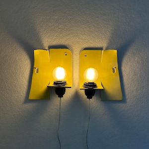 May include: A pair of wall-mounted sconces featuring bright yellow, flat, rectangular backplates with curved edges. Each sconce has a black socket holding a glowing, round, warm-toned light bulb, with a thin, clear power cord extending downward.