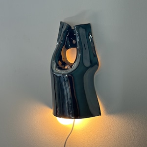 May include: A dark, glossy, ceramic wall sconce with a smooth, curved, abstract form. The fixture features a central cutout and emits a warm, glowing light from its base, casting a soft illumination downwards. A thin power cord hangs from the bottom.