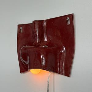 May include: A red ceramic wall light with a distinctive, textured form. The light has a warm yellow bulb. The power cable is visible, and the light is mounted on a white wall. A decorative piece for the home.