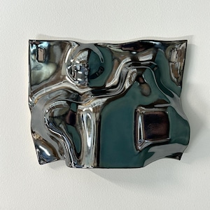 Pu&ograve; includere: Una placca da parete rettangolare in ceramica con una smaltatura argentata metallizzata altamente riflettente. La superficie presenta una texture irregolare, ondulata e simile a un liquido con curve e pieghe profonde. Due piccoli fori rettangolari sono posizionati in alto.