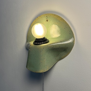 Puede incluir: Un aplique de pared con una pantalla de cer&aacute;mica curva de color verde p&aacute;lido con un patr&oacute;n de motas oscuras. La l&aacute;mpara incluye un casquillo circular negro que sostiene una bombilla esf&eacute;rica esmerilada encendida y un cable blanco fino.