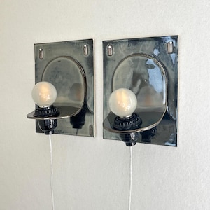 May include: Two wall-mounted sconces with a dark, glossy finish. Each sconce features a square backplate with an arched design and a round lightbulb. The sconces are connected to a power source via a white cord.