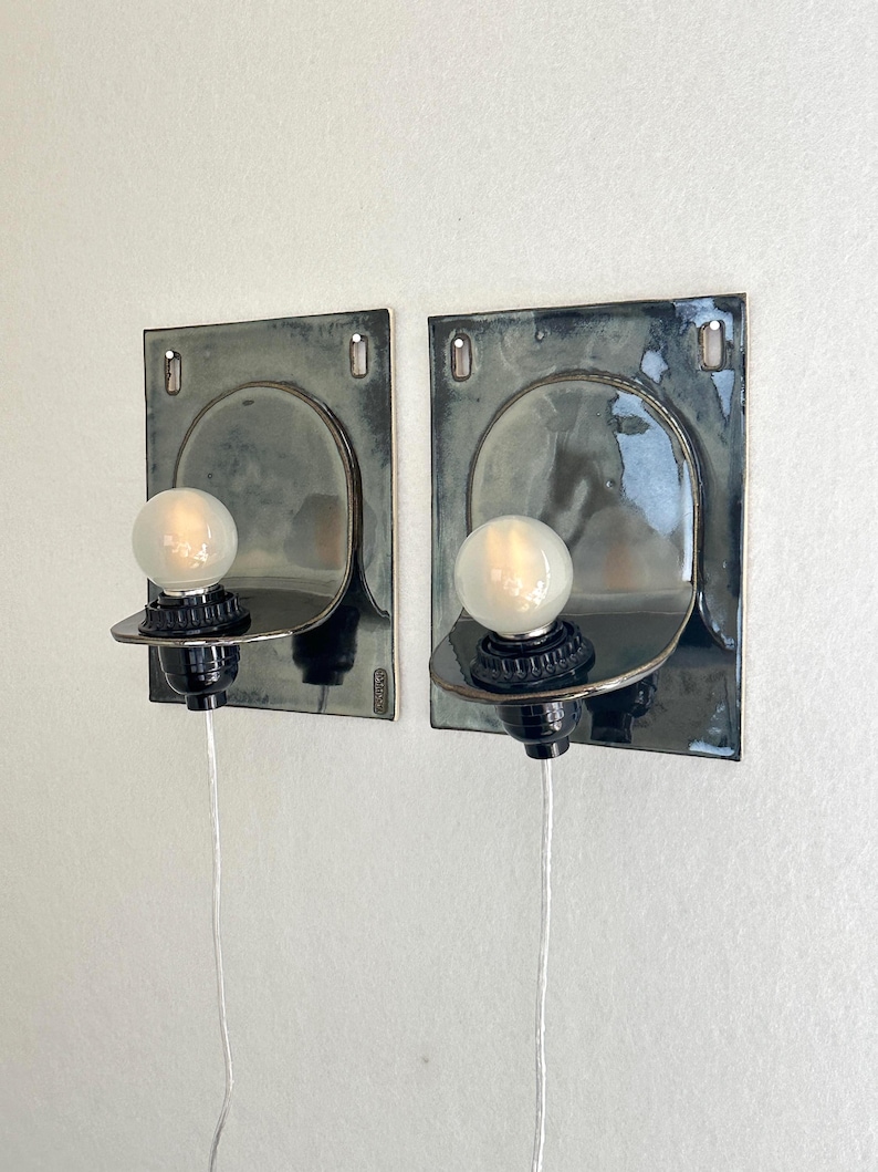 May include: Two wall-mounted sconces with a dark, glossy finish. Each sconce features a square backplate with an arched design and a round lightbulb. The sconces are connected to a power source via a white cord.