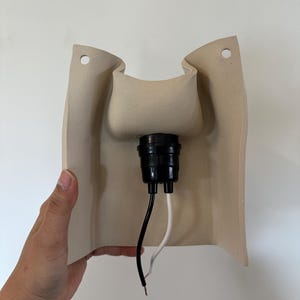 May include: A beige, curved wall-mounted light fixture base featuring a central black socket component. The structure includes two circular mounting holes at the top corners and two exposed electrical wires, one black and one white, extending downwards.