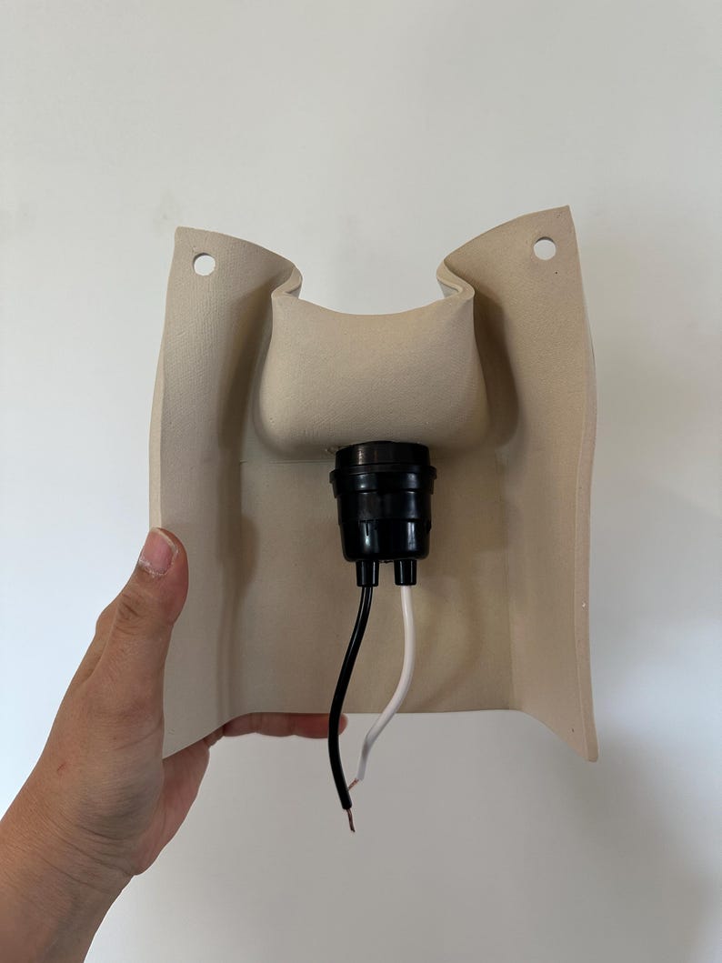 May include: A beige, curved wall-mounted light fixture base featuring a central black socket component. The structure includes two circular mounting holes at the top corners and two exposed electrical wires, one black and one white, extending downwards.
