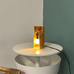 Puede incluir: Una l&aacute;mpara de mesa cil&iacute;ndrica con un acabado moteado en marr&oacute;n y bronceado, que presenta un dise&ntilde;o de corte curvo que revela una bombilla blanca brillante en su interior. La l&aacute;mpara tiene un cable negro y emite una luz c&aacute;lida y suave.