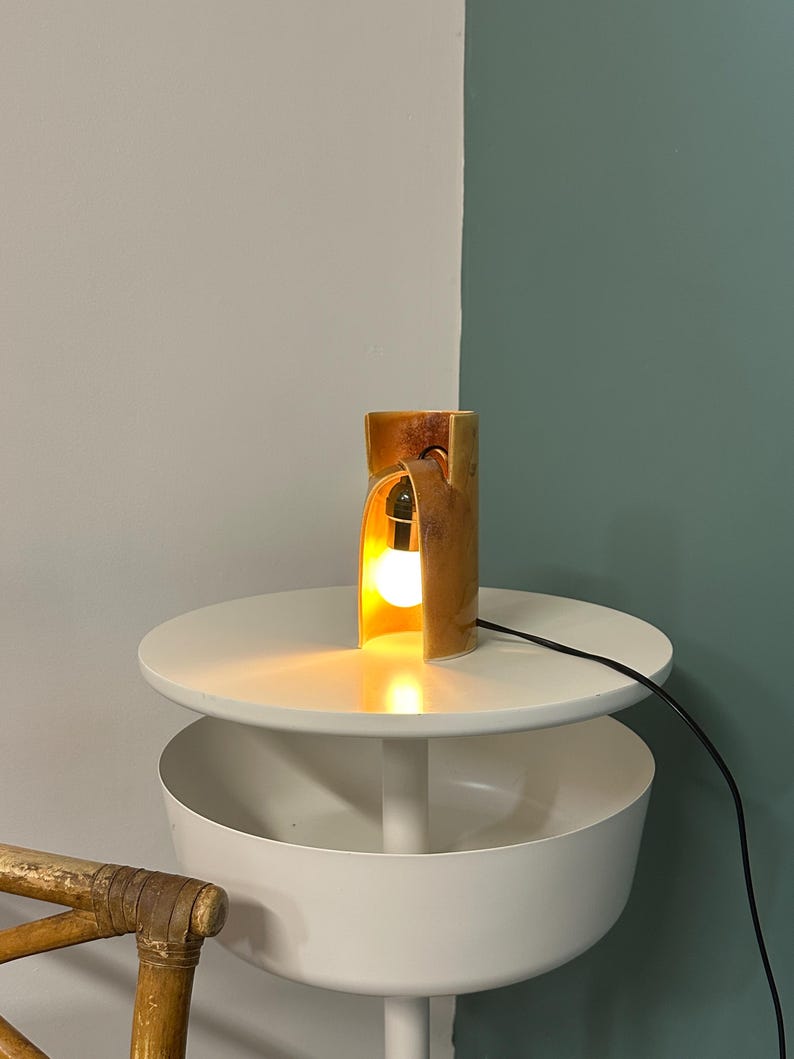 Puede incluir: Una l&aacute;mpara de mesa cil&iacute;ndrica con un acabado moteado en marr&oacute;n y bronceado, que presenta un dise&ntilde;o de corte curvo que revela una bombilla blanca brillante en su interior. La l&aacute;mpara tiene un cable negro y emite una luz c&aacute;lida y suave.