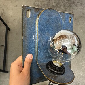 May include: A wall-mounted light fixture featuring a rectangular blue ceramic backplate with rounded edges and two mounting holes. A circular ceramic shelf extends from the center, holding a black socket with a large, reflective silver globe bulb.