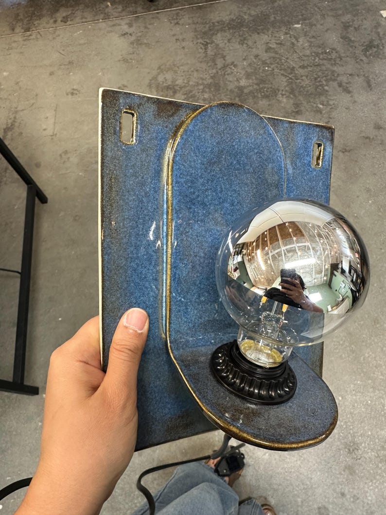 May include: A wall-mounted light fixture featuring a rectangular blue ceramic backplate with rounded edges and two mounting holes. A circular ceramic shelf extends from the center, holding a black socket with a large, reflective silver globe bulb.