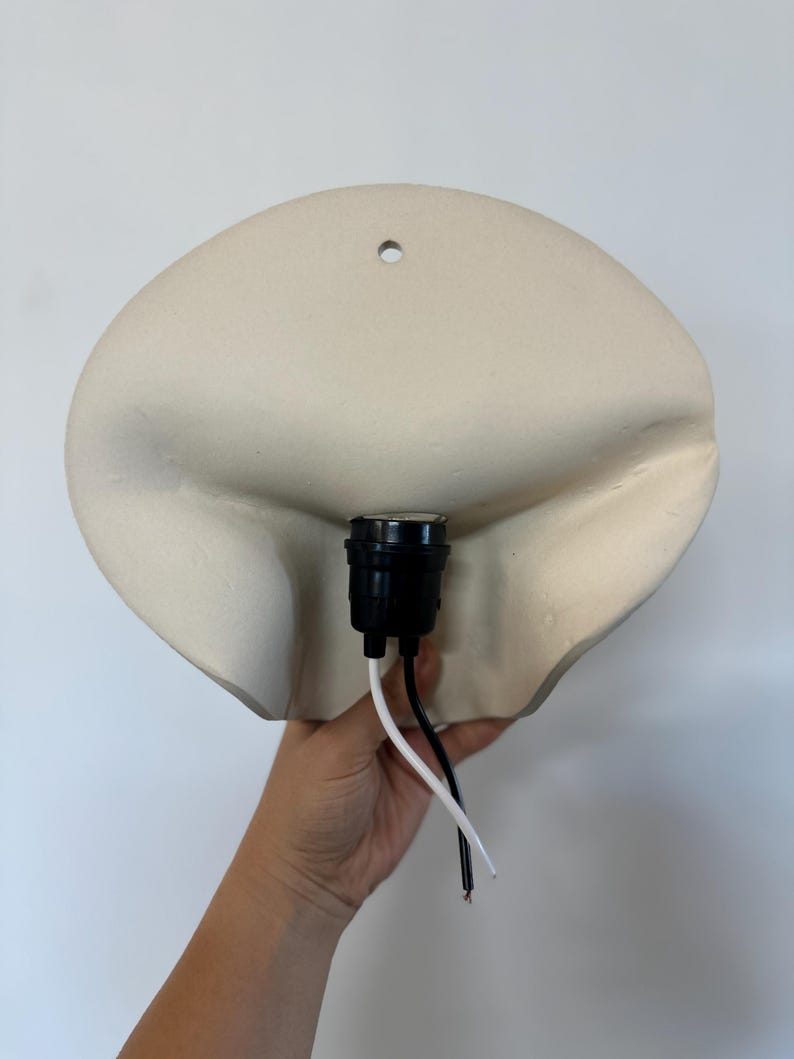May include: A wall-mounted light fixture featuring a matte off-white ceramic shade with a soft, organic, folded shape. The center includes a black plastic light socket with one white wire and one black wire extending downward for electrical connection.