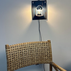 May include: A wall-mounted light fixture featuring a dark rectangular metal backplate and a single clear glass bulb. Below the light, the upper portion of a chair with a woven light brown cane backrest and a wooden frame is visible.