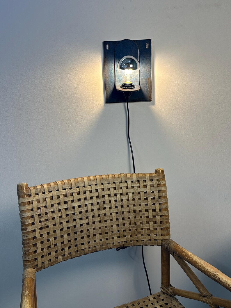 May include: A wall-mounted light fixture featuring a dark rectangular metal backplate and a single clear glass bulb. Below the light, the upper portion of a chair with a woven light brown cane backrest and a wooden frame is visible.