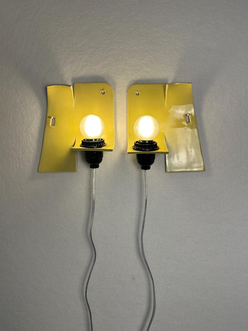 May include: Two yellow ceramic wall sconces with an irregular, folded shape. Each fixture features a black socket holding a glowing, round white light bulb. A thin, silver-colored electrical cord hangs downward from the base of each lamp.
