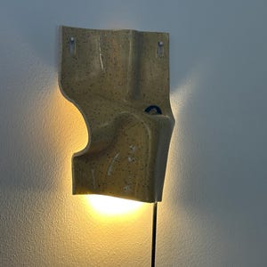 K&ouml;nnte beinhalten: Eine Wandleuchte aus Keramik mit gl&auml;nzend gelber Oberfl&auml;che und dunklen Sprenkeln. Die Leuchte hat eine unregelm&auml;&szlig;ige, gewellte Form mit einem kleinen blauen kreisf&ouml;rmigen Akzent und strahlt durch ein schwarzes Kabel warmes Licht nach unten ab.