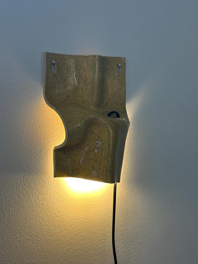 K&ouml;nnte beinhalten: Eine Wandleuchte aus Keramik mit gl&auml;nzend gelber Oberfl&auml;che und dunklen Sprenkeln. Die Leuchte hat eine unregelm&auml;&szlig;ige, gewellte Form mit einem kleinen blauen kreisf&ouml;rmigen Akzent und strahlt durch ein schwarzes Kabel warmes Licht nach unten ab.
