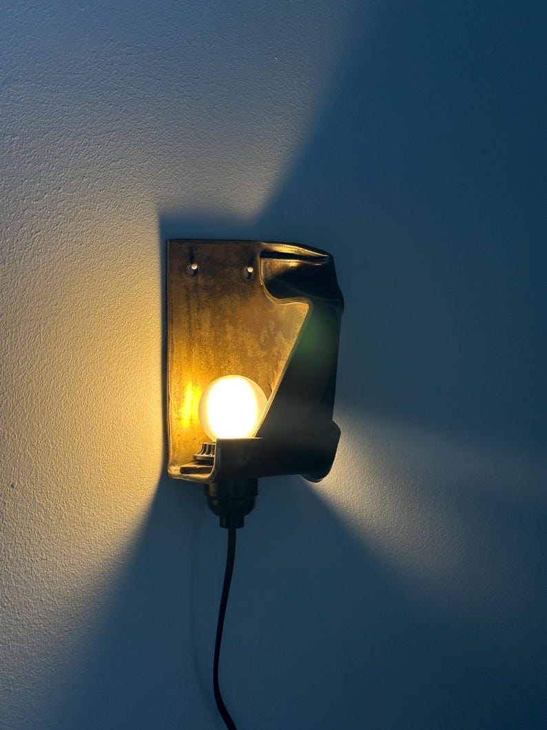 May include: A wall-mounted sconce featuring a dark, curved metal housing with a textured finish. A single round, glowing light bulb is positioned within the structure, casting a warm yellow light against the wall and creating a distinct shadow.