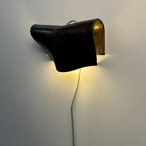 May include: A wall-mounted lamp featuring a curved, dark brown leather shade with a metallic gold interior finish. The light emits a warm glow from beneath the shade, and a thin, silver-coloured electrical cord hangs vertically from the base.