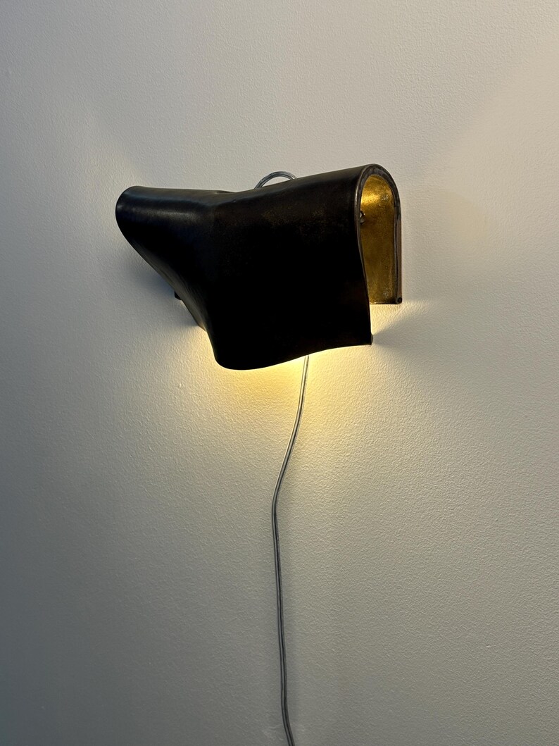 May include: A wall-mounted lamp featuring a curved, dark brown leather shade with a metallic gold interior finish. The light emits a warm glow from beneath the shade, and a thin, silver-coloured electrical cord hangs vertically from the base.