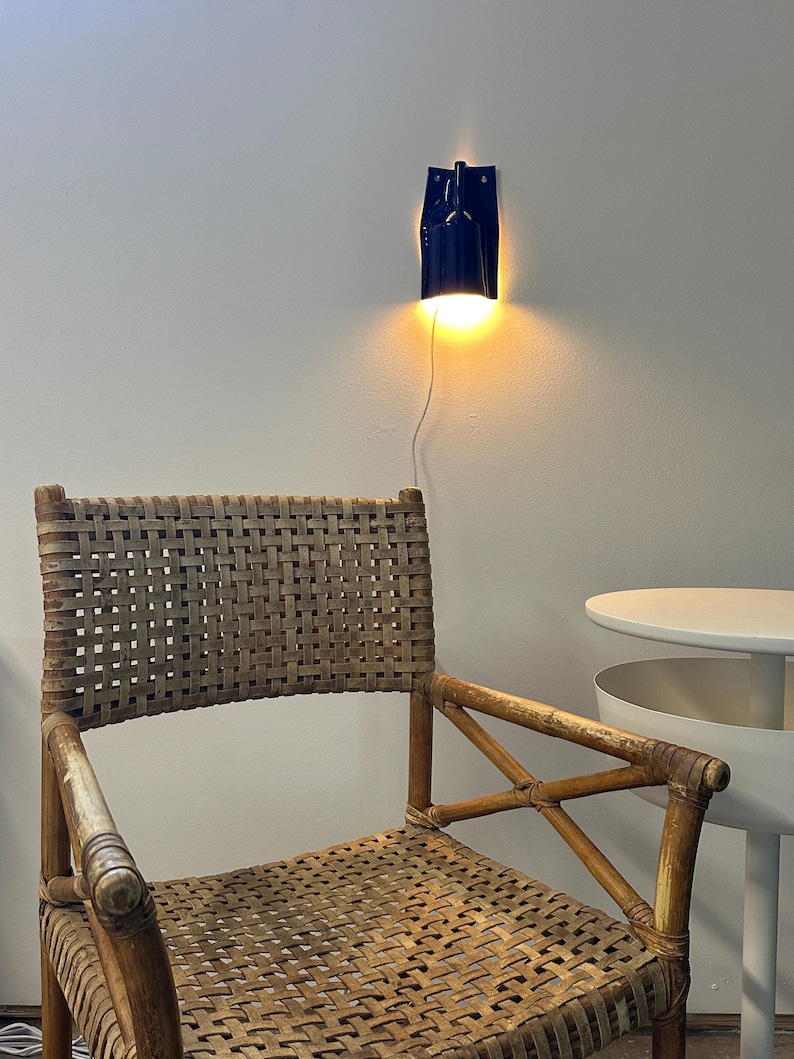 Puede incluir: Un sill&oacute;n de rat&aacute;n trenzado con acabado en marr&oacute;n claro natural y estructura de refuerzo en cruz. Sobre la silla, una l&aacute;mpara de pared azul oscuro emite una luz c&aacute;lida y brillante. A la derecha, una mesa auxiliar redonda blanca con estante.