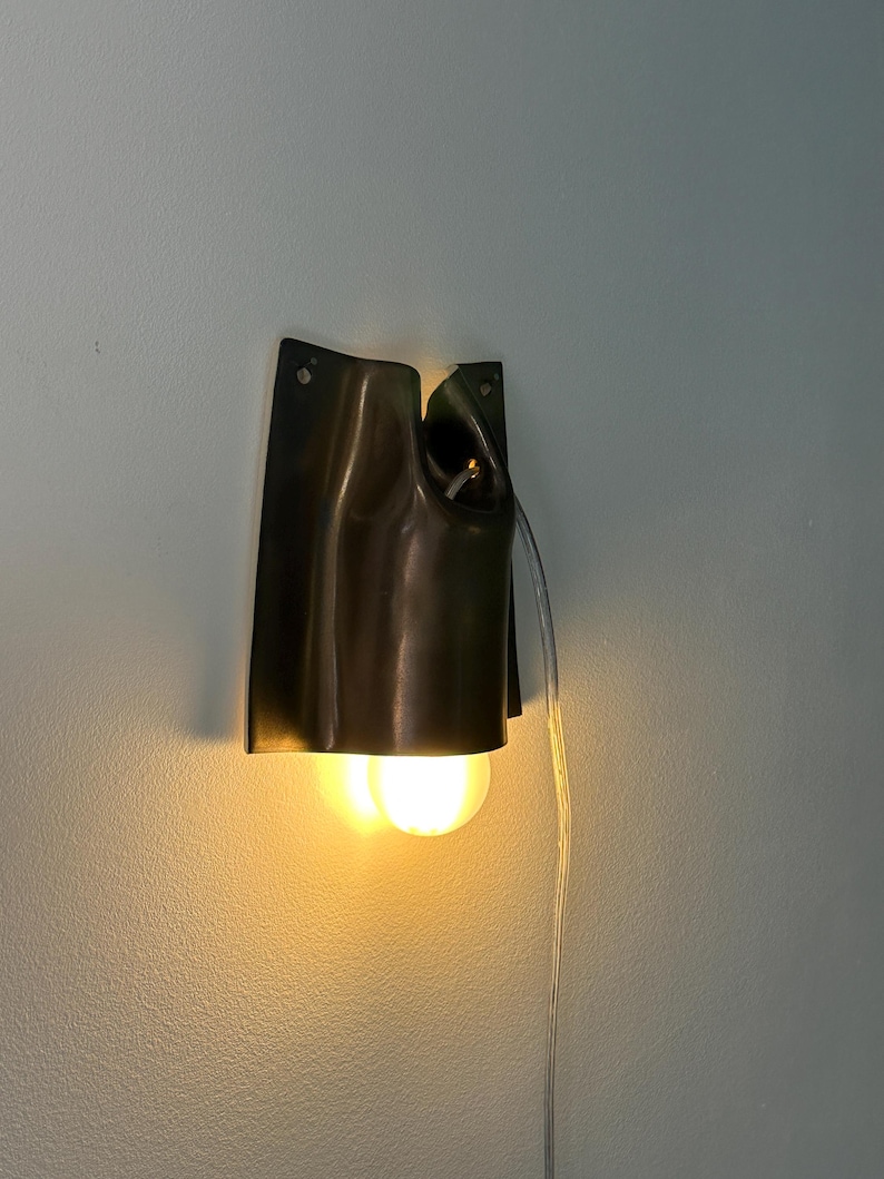 May include: A dark brown, curved metal wall sconce with a smooth, folded silhouette. The fixture features a single exposed light bulb emitting a warm, yellow glow from the bottom opening. A clear electrical cord hangs down from the side of the unit.