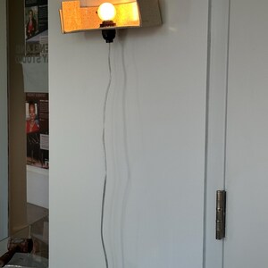 May include: A wall-mounted light fixture with a rectangular, textured shade. The light bulb is round and emits a warm glow. A black socket and a clear cord with an inline switch complete the design. The fixture is mounted on a white wall.
