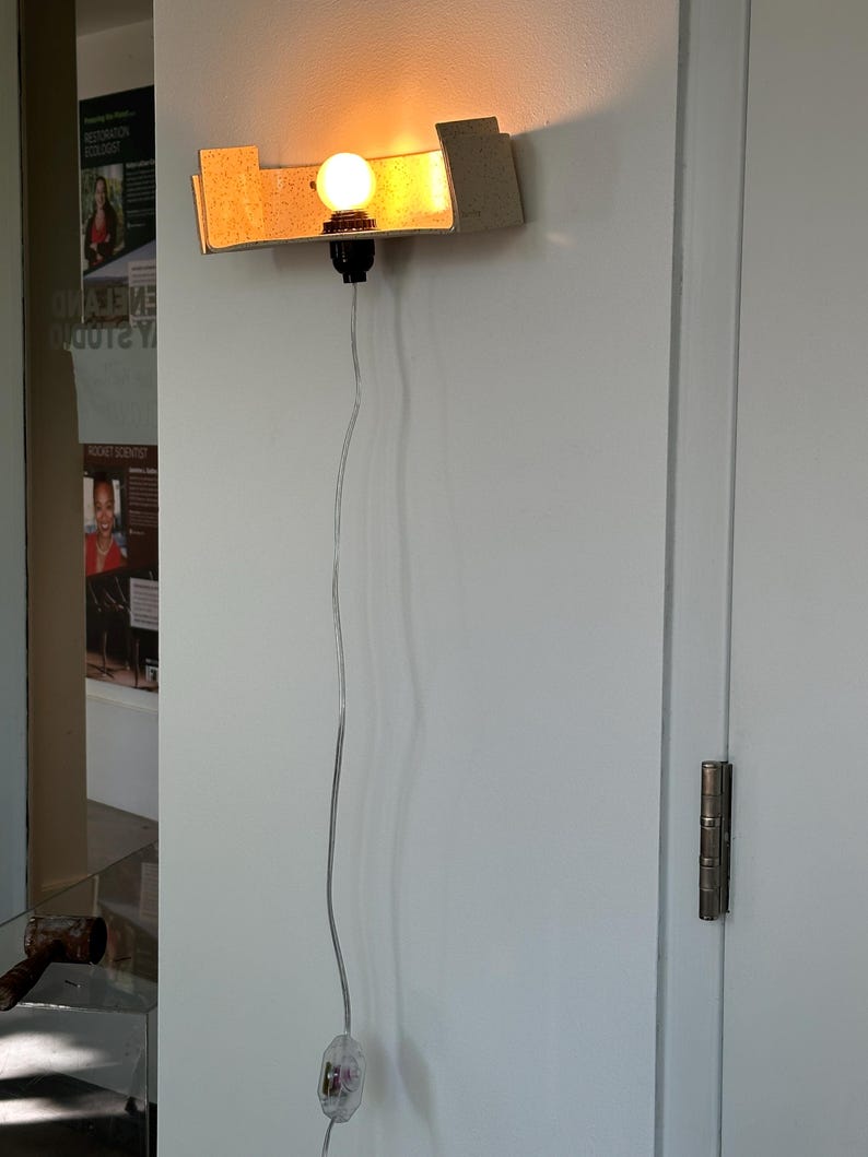 May include: A wall-mounted light fixture with a rectangular, textured shade. The light bulb is round and emits a warm glow. A black socket and a clear cord with an inline switch complete the design. The fixture is mounted on a white wall.