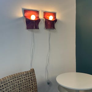 May include: Two wall-mounted sconces featuring a curved, rectangular design with a gradient of deep red to dark purple. Each fixture holds a single glowing round light bulb and includes a white power cord extending downward from the base.