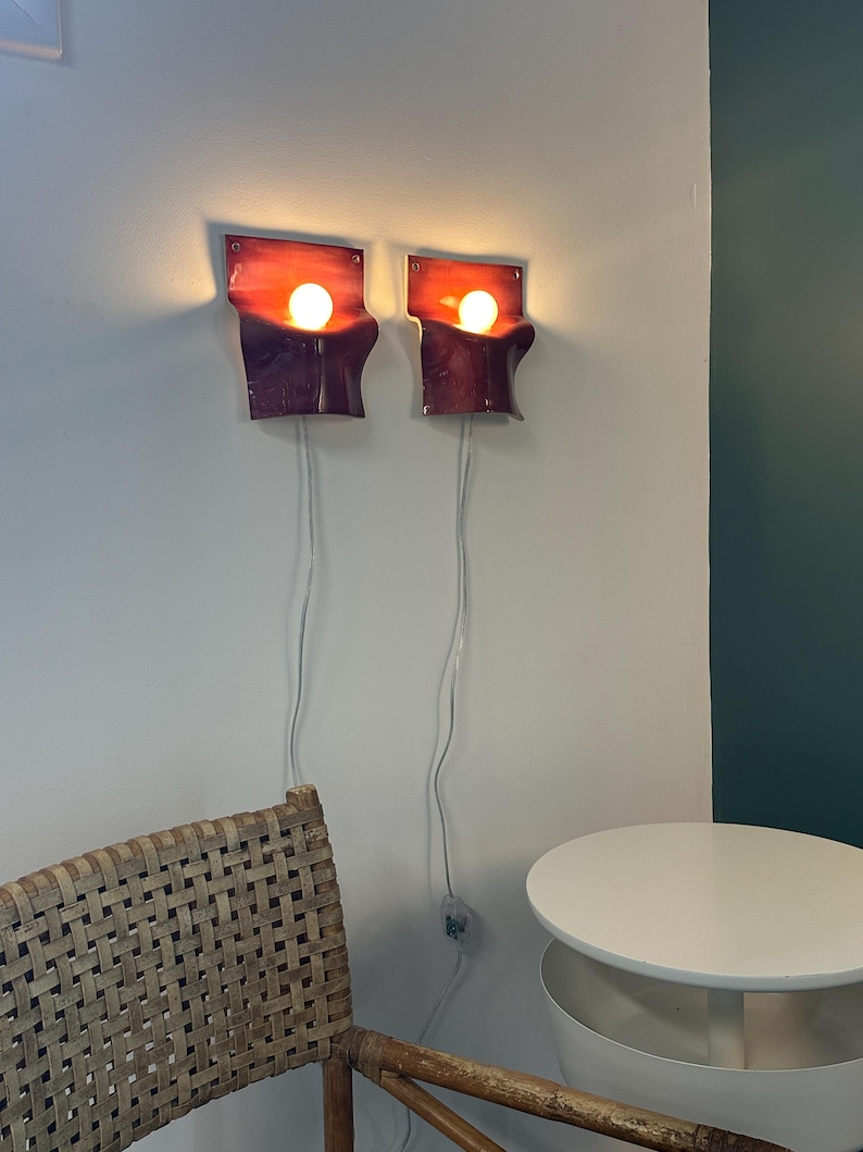 May include: Two wall-mounted sconces featuring a curved, rectangular design with a gradient of deep red to dark purple. Each fixture holds a single glowing round light bulb and includes a white power cord extending downward from the base.